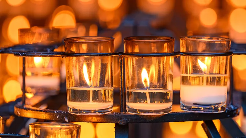 Lit votive candles in glass holders sit on a black metal rack at the Grotto of Our Lady of Lourdes.
