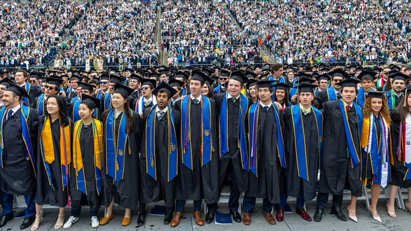 A diverse group of University of Notre Dame graduates in black caps and gowns with blue, gold, and green stoles stand and smile together. Behind them, a massive crowd of spectators fills the tiered seating of Notre Dame Stadium.