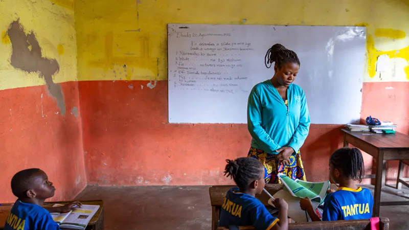 A woman in a teal jacket and colorful skirt stands in a classroom with orange and yellow walls.