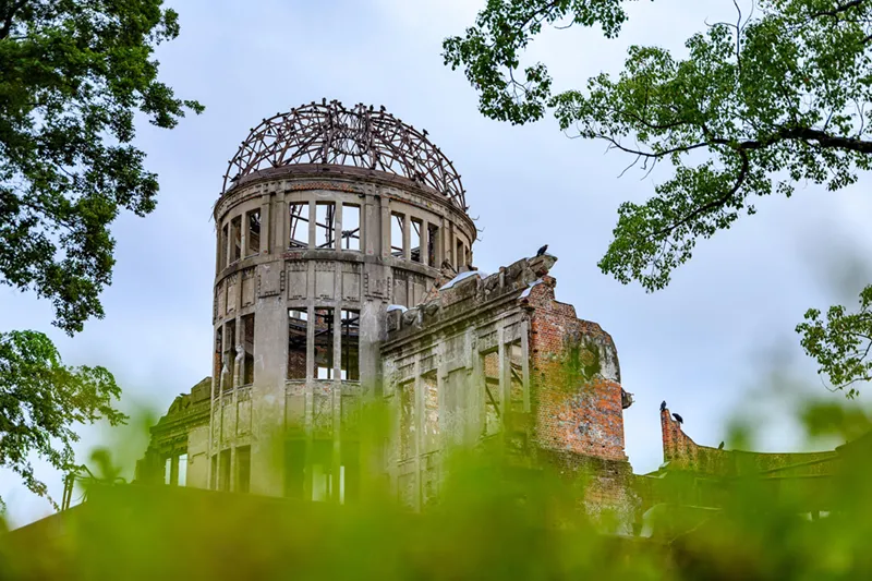 The skeletal dome and ruined brick walls of the Hiroshima Peace Memorial, viewed through green foliage.