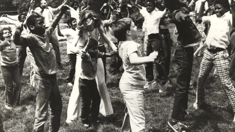 A diverse group of children and teens dance energetically outdoors on a grass field in this vintage black-and-white photo, arms raised and bodies in motion, wearing 1970s-era casual clothing.