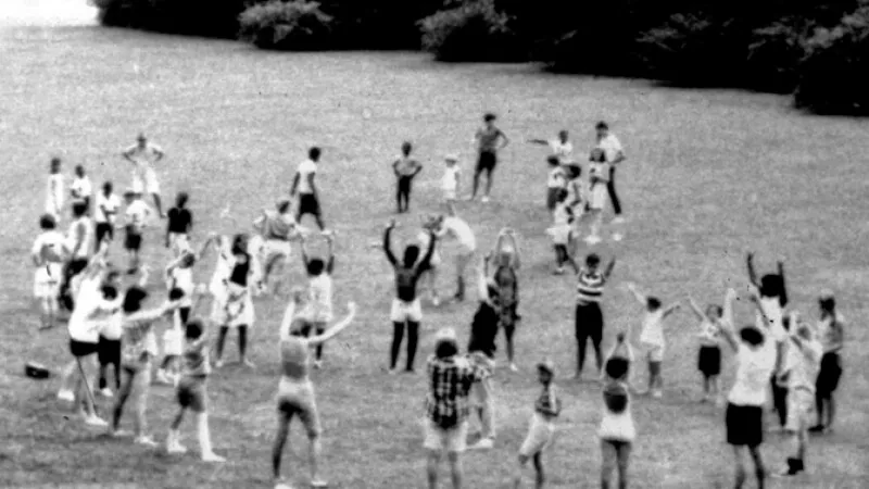 An aerial black-and-white view of a large group of children spread across an open grass field, arms raised during an outdoor group exercise or activity session.