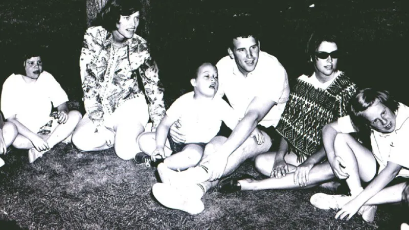 Five children and two young adults sit together on the ground near a tree in a vintage black-and-white photo, with a young man in a white shirt interacting warmly with a laughing child beside him.