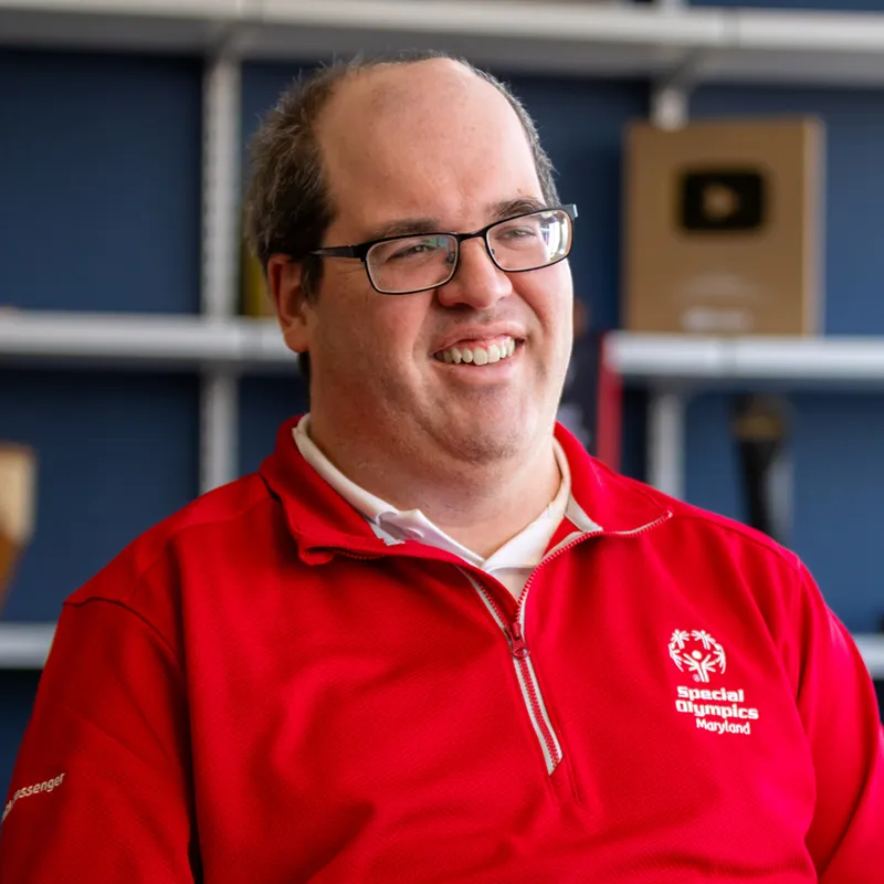 A smiling man with glasses and short brown hair is wearing a red quarter-zip pullover. The pullover features the Special Olympics Maryland logo on the chest and the word messenger on the sleeve. In the background are shelves with various awards.