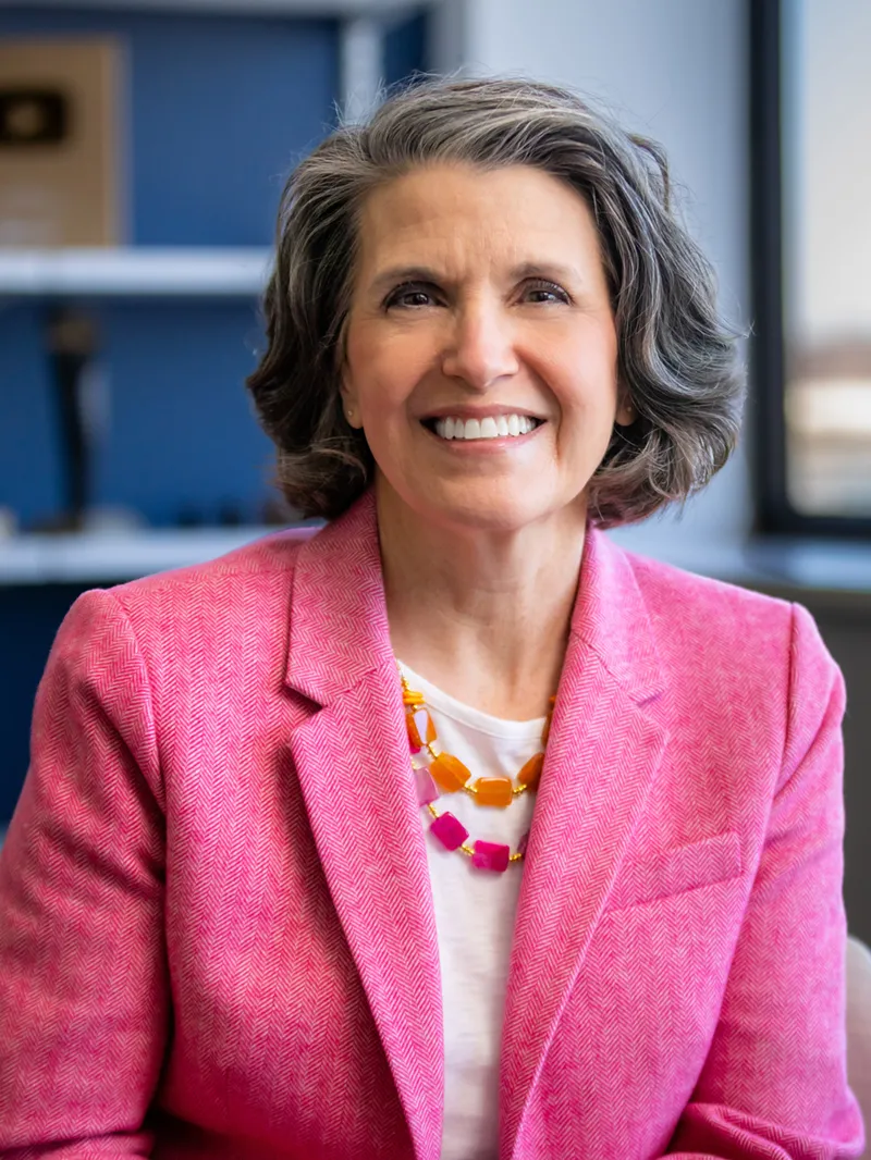Smiling woman with short gray hair wearing a pink herringbone blazer and a colorful orange and pink gemstone necklace, seated in a bright office environment.