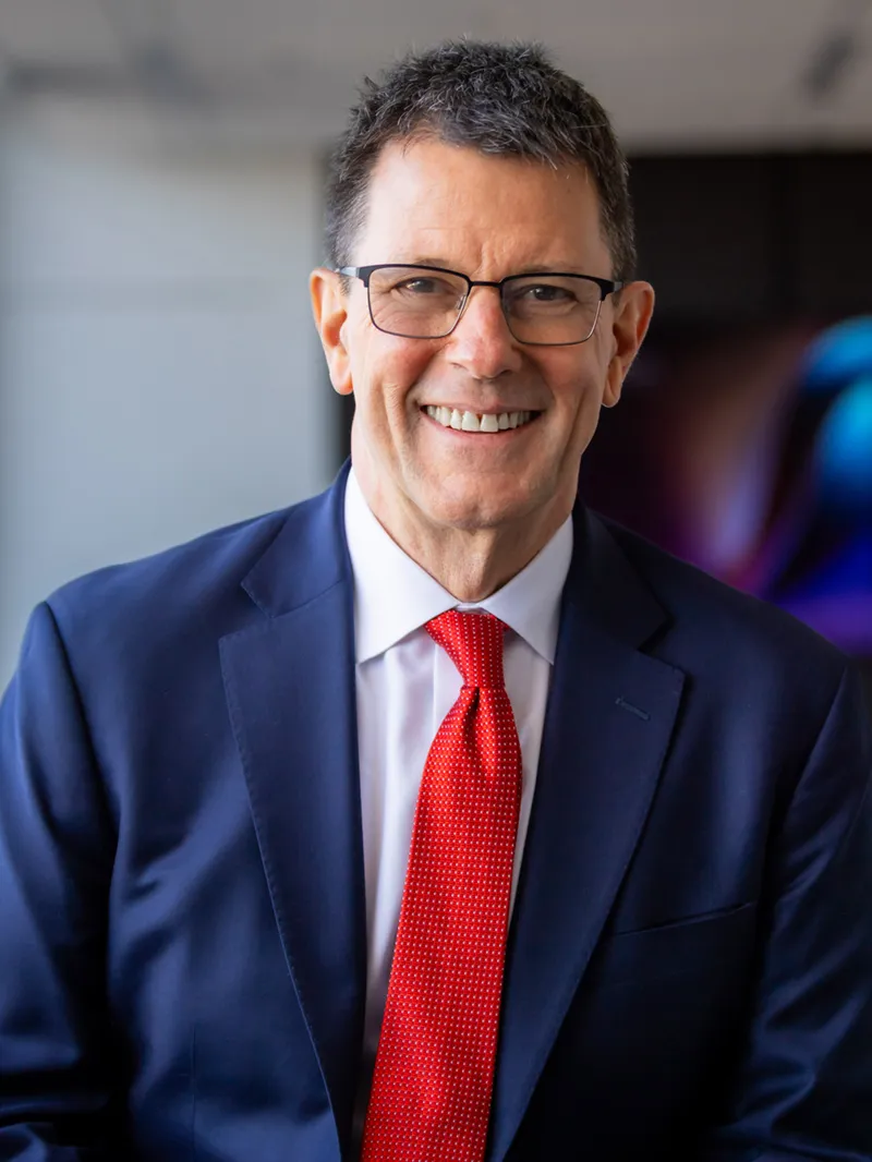 Smiling man with short dark hair and rectangular glasses wearing a navy blazer, white dress shirt, and red tie, with a blurred colorful background behind him.