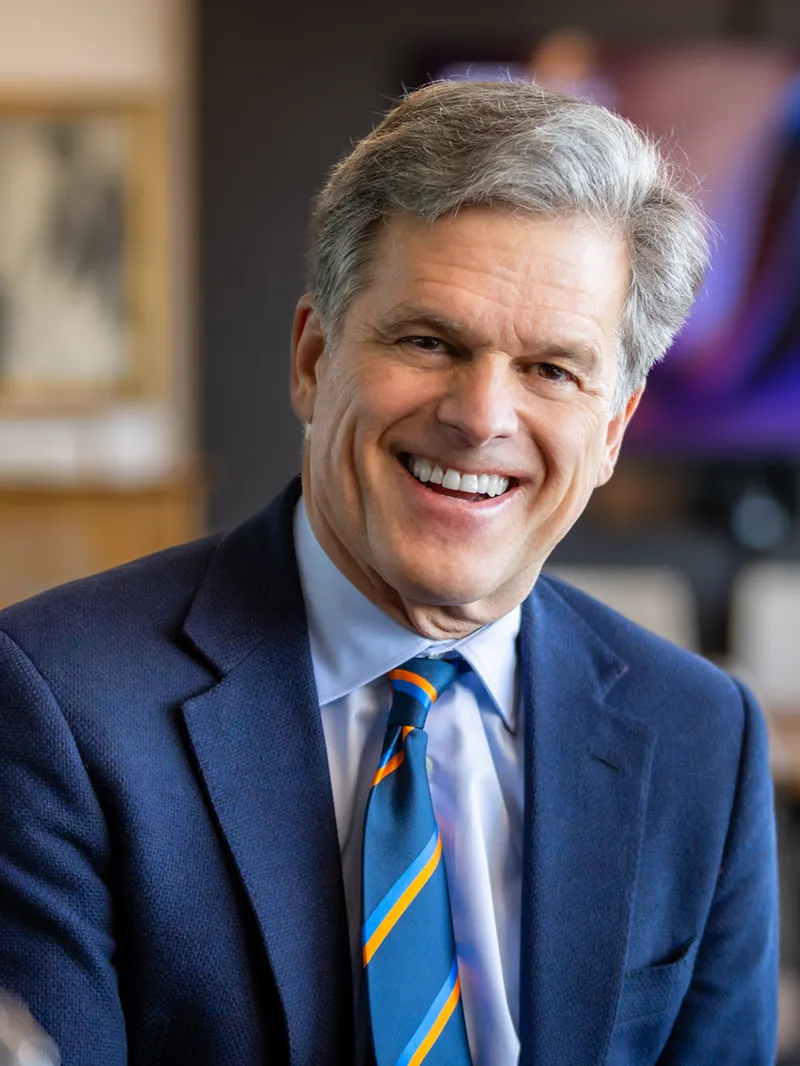 Smiling man with gray hair wearing a navy blazer, light blue shirt, and blue striped tie with orange accents, with a blurred warm-toned interior space visible behind him.