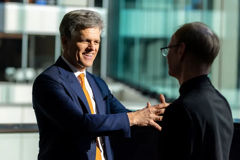 A smiling man in a navy suit and orange tie gestures warmly while speaking with a person in a black clerical suit in a modern glass-walled building interior.
