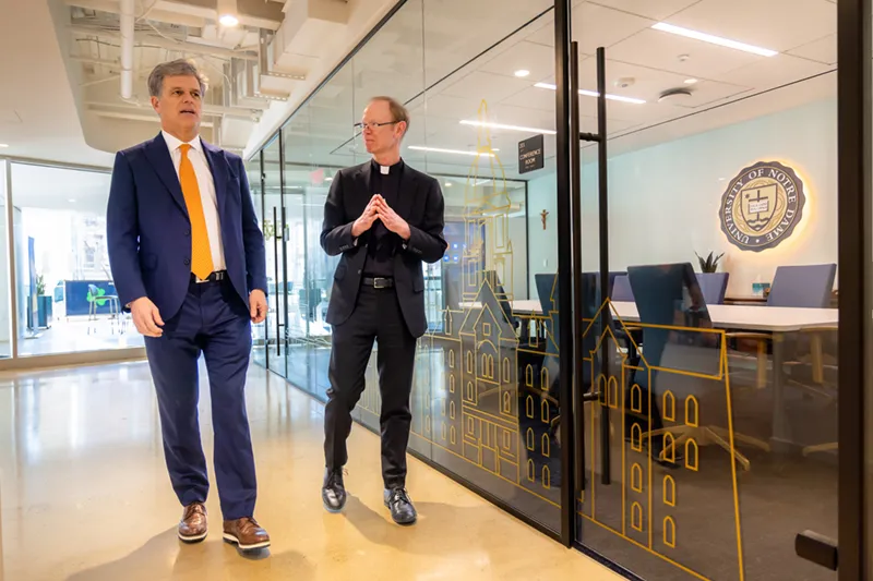 A man in a navy suit and orange tie walks alongside a Catholic priest in a black clerical suit past a glass partition etched with a gold Notre Dame campus skyline illustration and the University seal.