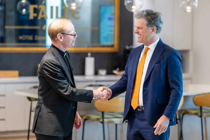 A Catholic priest in a black clerical suit shakes hands with a smiling man in a navy suit and orange tie inside a modern space with a gold 'Fáilte' sign visible in the background.