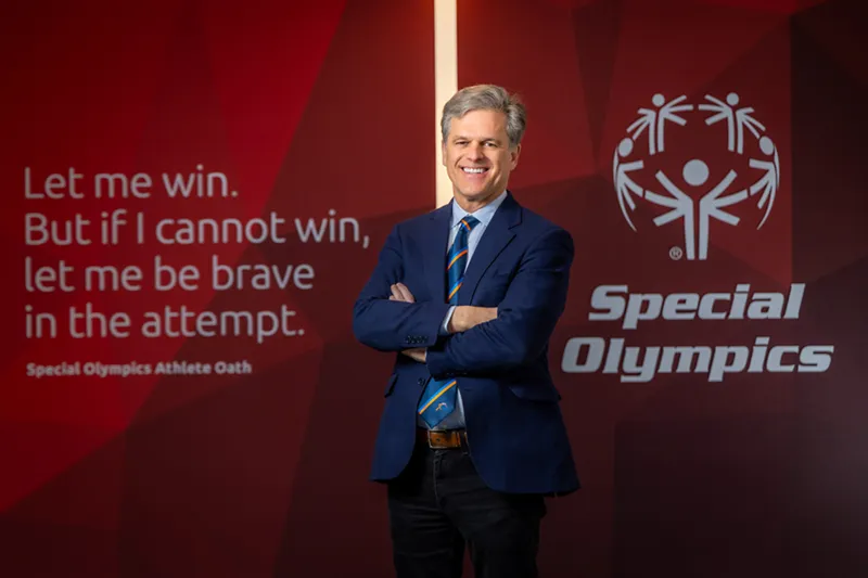 Timothy Shriver smiles with his arms crossed while wearing a navy blue suit and a striped orange and blue tie. He stands in front of a blurred maroon background featuring the Special Olympics logo and text.