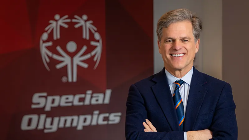 Smiling man with gray hair, arms crossed, wearing a navy blazer, light blue shirt, and blue striped tie with orange accents, standing before a large red Special Olympics logo and wordmark.