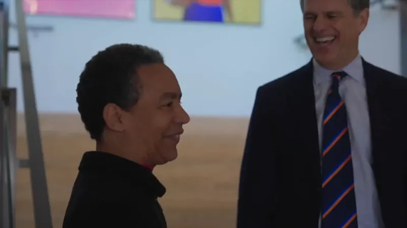 Two people share a warm, laughing exchange indoors — one in a black turtleneck facing left in profile, the other in a navy suit with a blue and orange striped tie, with colorful artwork visible on the wall behind them.