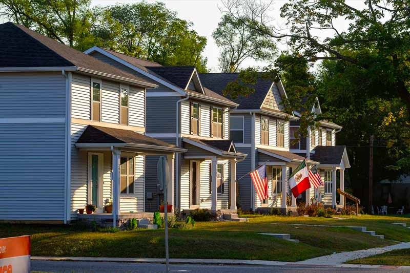 A street block of four two-story, newly constructed homes. Two of the homes fly an American flag. A home in between the two fly a Mexican flag.