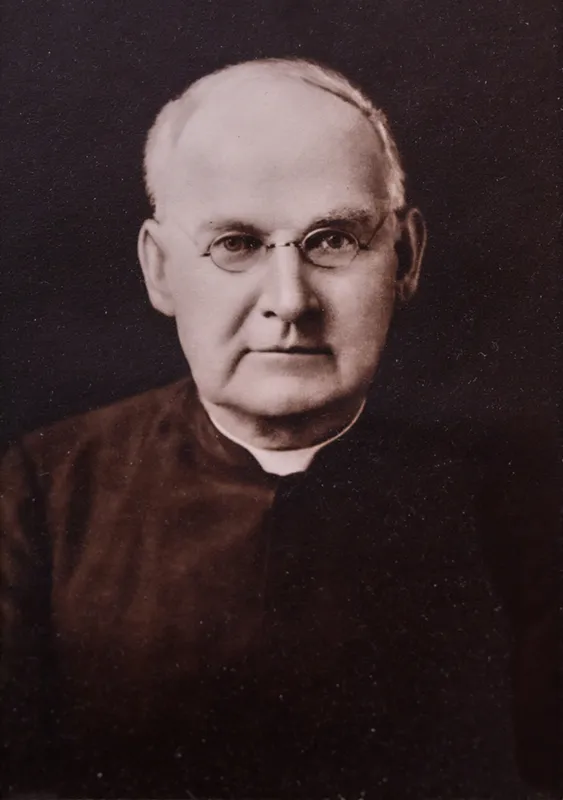 Historical portrait of Brother Columba O'Neill, CSC, wearing a dark religious habit and white collar. The older man has thinning hair and round wire-frame glasses with a serious expression against a dark background.