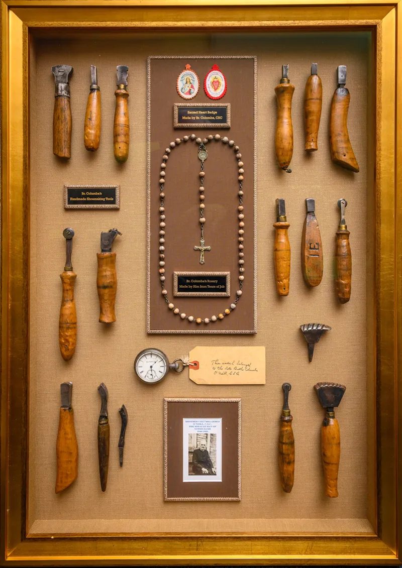 A wooden display case with a gold frame contains a collection of Brother Columba's cobbler tools and personal devotional items arranged on tan fabric. Along the left side are six wooden-handled tools with metal heads of varying shapes, used for shoe repair. On the right side are six more similar tools. At the center is a beaded rosary with a silver cross and medal hanging from a brown backing with a small brass nameplate reading 'St. Columba's Rosary made by Fr. Columba, C.S.C.' Above the rosary are two oval religious medals with red backgrounds. Below is another small brass plaque. At the bottom center is a vintage pocket watch with white face and Roman numerals, a handwritten note in cursive script, and a framed sepia-toned photograph of an elderly man in religious robes. Additional items include wire brushes and what appear to be burnishing tools.