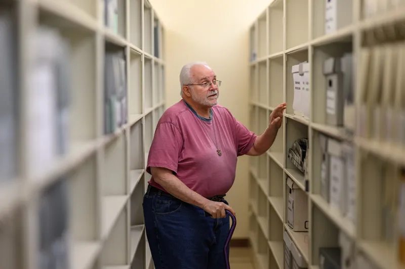 An older man with white hair, a beard, and glasses reaches for a box in an archive aisle. He wears a pink t-shirt and a cross necklace while holding a purple cane. Rows of tall, grey metal shelves filled with boxes extend into the background.