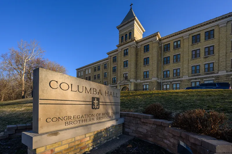 A stone sign for Columba Hall, Congregation of Holy Cross Brothers Residence, stands in the foreground. In the background is the multi-story yellow brick Columba Hall at the University of Notre Dame, featuring a central tower topped with a cross.