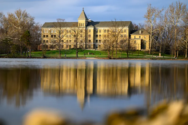 Columba Hall at the University of Notre Dame, a yellow brick building with a central tower, reflected in the still water of St. Mary's Lake. Bare trees and a walking path with small figures border the green lawn in front of the hall.