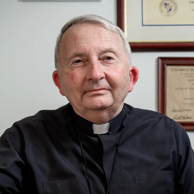 An older man with white hair wearing black clerical robes with a white collar sits in an office. He has a serious, composed expression and is photographed from the shoulders up.
