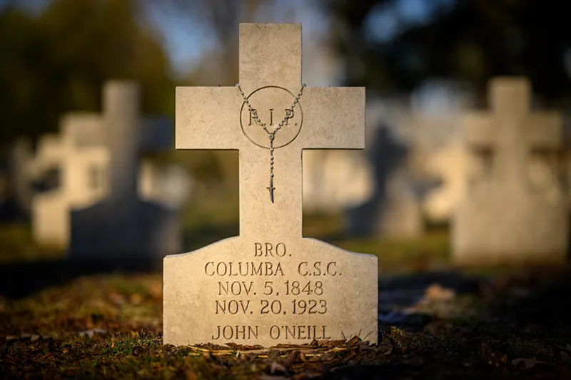 Brother Columba's grave at Holy Cross Cemetery on the Notre Dame campus, featuring a white marble cross with R.I.P. inscribed in a circle at its center. The base reads: BRO. COLUMBA C.S.C. NOV. 5, 1848 NOV. 20, 1923 JOHN O'NEILL. The grave is surrounded by other white crosses in soft focus, with grass and natural ground cover visible. The image conveys a sense of reverence and peaceful remembrance.