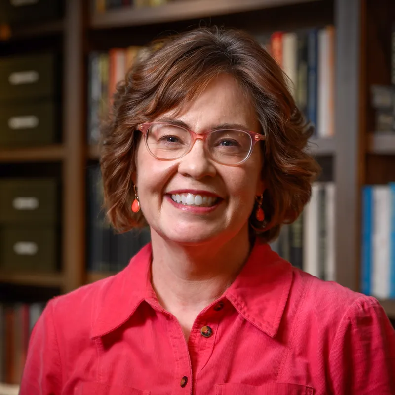 A woman with shoulder-length reddish-brown hair and glasses smiles warmly at the camera while wearing a bright red button-up shirt. She stands in front of wooden bookshelves filled with books in various colors, with filing cabinets visible in the soft-focused background.