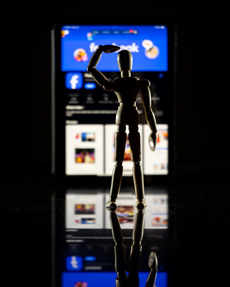 A wooden artist's mannequin stands on a dark, reflective surface, facing a glowing tablet displaying the Facebook interface. The mannequin has one arm raised to its head as if peering forward into the light against a black background.