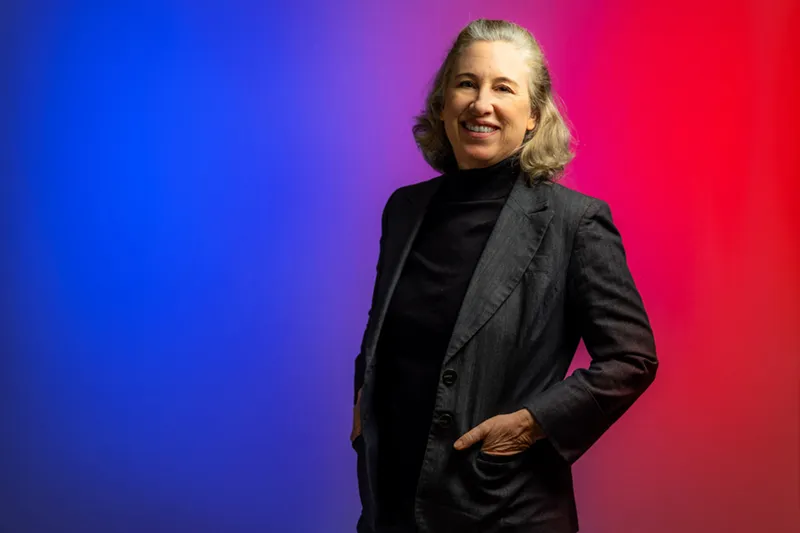 Lisa Schirch smiles in a professional headshot. She has wavy grey-blonde hair and wears a dark grey blazer over a black turtleneck sweater, posed against a gradient blue to red background.