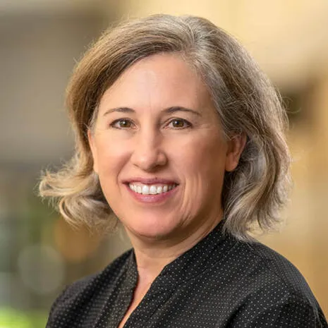 Lisa Schirch smiles in a professional headshot. She has short, wavy grey-blonde hair and brown eyes, and is wearing a black blouse with a white micro-dot pattern against a soft-focus, warm-toned background.