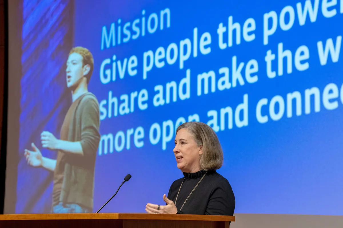 A woman with grey-blonde hair speaks at a podium in front of a large blue screen. The screen displays a photo of Mark Zuckerberg and text about the power to share and connect. She wears a black top and gestures with her hands while speaking.