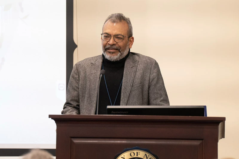A man with a grey beard and glasses, wearing a black turtleneck and grey blazer, stands behind a dark wood lectern. He looks forward with a slight smile while speaking at a microphone during an event at the University of Notre Dame.