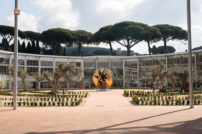 The bronze sculpture Desert Rose by Arnaldo Pomodoro is placed at the center of the ecological greenhouse at the Gardens of Castel Gandolfo. The curved glass structure&mdash;covered with solar panels&mdash;frames the work within a light-filled, sustainable environment. Stone pathways and carefully trimmed hedges unfold beneath a canopy of Mediterranean pines.