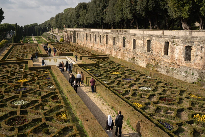 Visitors walk along a gravel path through the ornate, geometric knot gardens of the Pontifical Villas of Castel Gandolfo. High stone walls and a line of lush trees overlook the intricate flowerbeds and a central reflecting pool.
