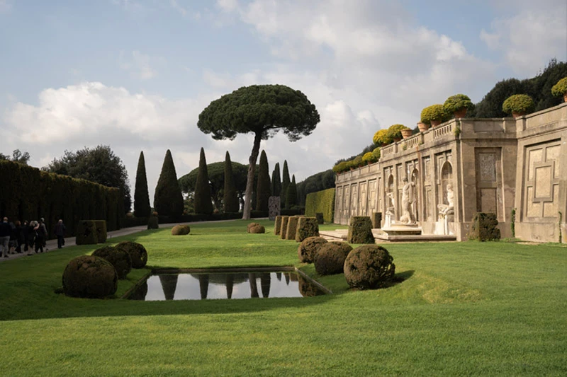 A stone fountain with classical statues borders a manicured lawn and reflecting pool at the Gardens of Castel Gandolfo. Distinctive umbrella pines and cypress trees rise against a soft blue sky behind a row of terracotta potted plants.