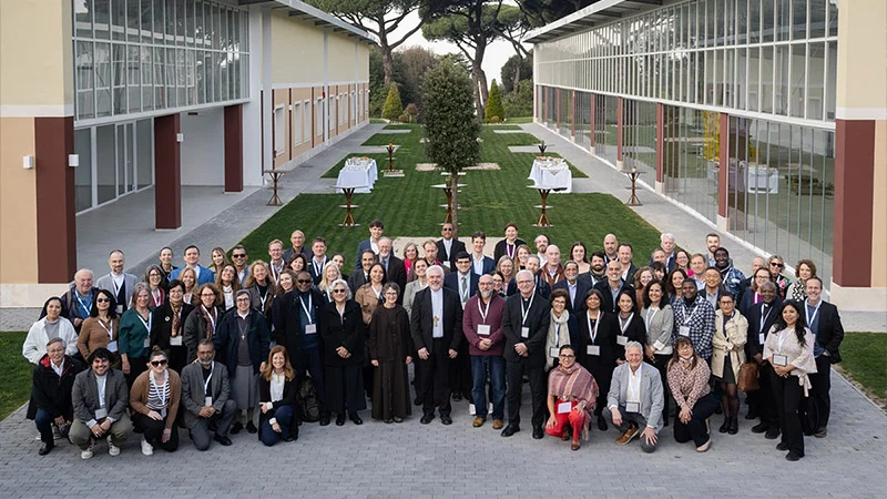 Diverse group of 70+ people, including clergy, smiling and wearing lanyards, pose in an outdoor courtyard with modern buildings.