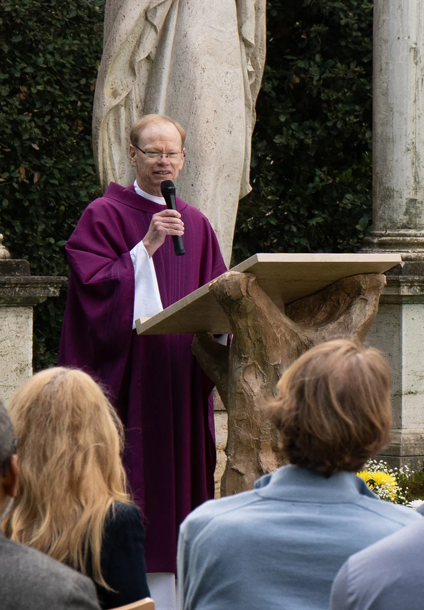 A white male priest wearing purple liturgical vestments, speaks into a microphone at a rustic stone lectern during an outdoor Mass. A statue of the Virgin Mary stands behind him as an audience listens from the foreground.