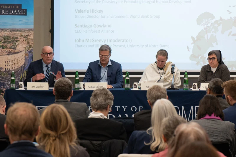 John McGreevy speaks at a microphone while seated at a blue table with three other panelists. A University of Notre Dame banner stands to the left, and a large projection screen displays the speakers&rsquo; names behind them.