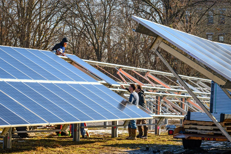 Workers in casual clothing and boots install large blue solar panels onto metal frames in an open field. Bare trees and a clear sky are visible in the background of the construction site.
