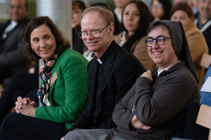 A woman in a green blazer, a priest in a black clerical suit, and a nun in a brown habit sit together and smile during an event. They are seated in an audience with several other people blurred in the background.