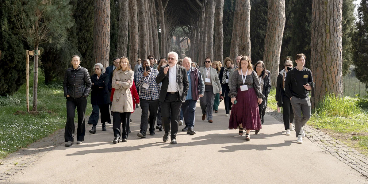 A diverse group of people walk down a paved path lined with tall, slender trees. A man in a black suit and white shirt leads the group. Many participants wear lanyards and casual professional attire under a bright, overcast sky.