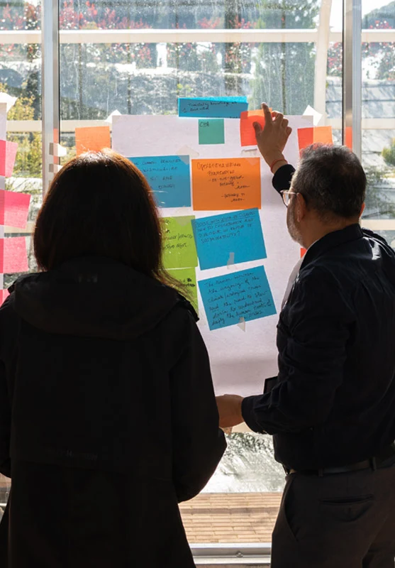 From behind, a woman and a man in a black button-down and glasses review colorful sticky notes on a glass window. Notes contain text about climate crisis and sustainability. Bright greenery is visible through the glass.
