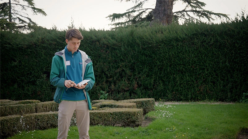 A young man in a green jacket and tan pants stands in a grassy garden, looking down as he writes in a small notebook. A tall, manicured hedge and large evergreen trees provide a lush green backdrop under a bright, overcast sky.