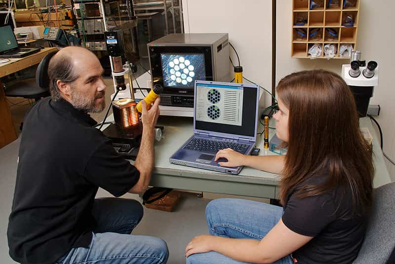 A	teacher and a student have a conversation sitting at a computer desk.