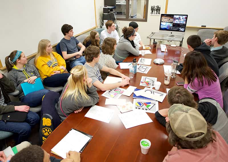 Students gather around a large, rectangular table with a computer sitting at the front and	four other people on screen for a video conference.