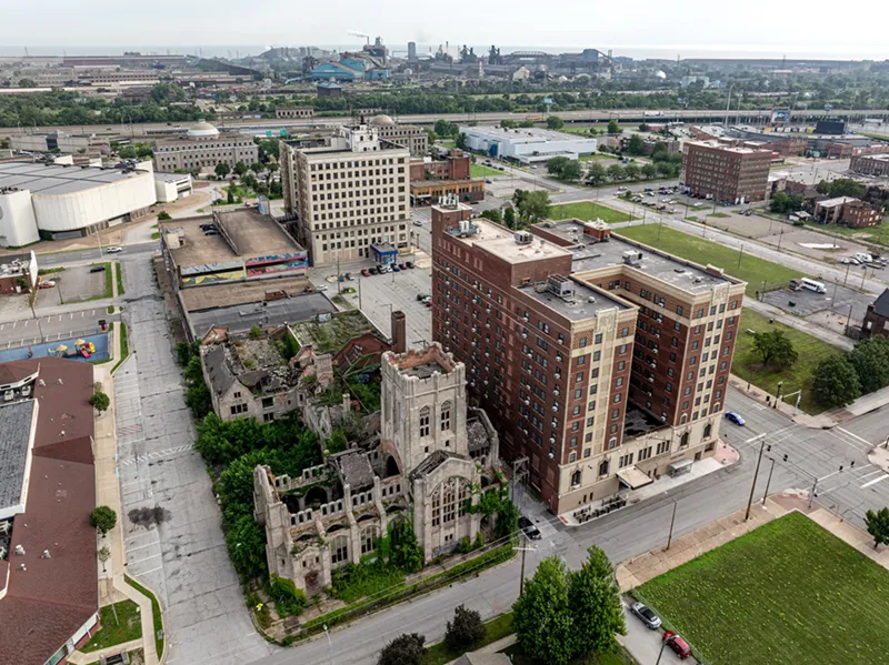 Aerial view of an abandoned, ivy-covered stone church ruin next to a tall red brick hotel and other buildings with U.S. Steel operations on the skyline in Gary, Indiana.