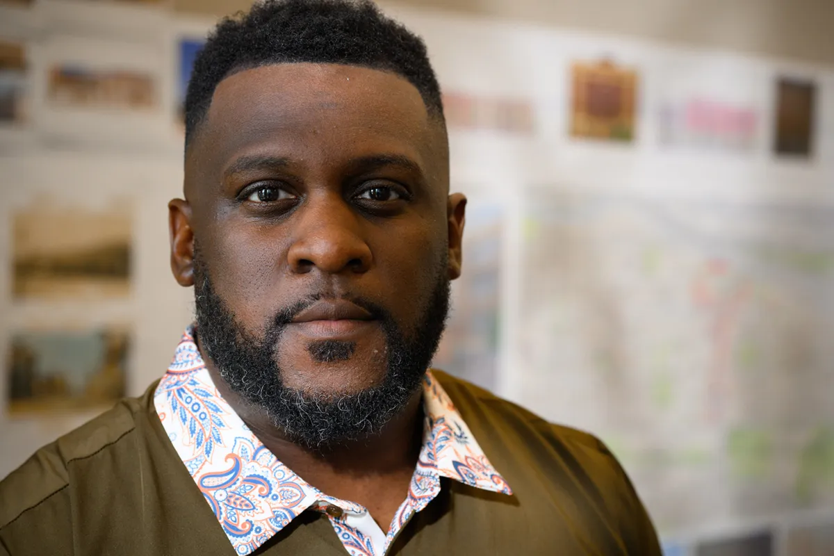 A man with a closely trimmed haircut and beard, wearing a green shirt with a patterned collar, looks intently at the camera.