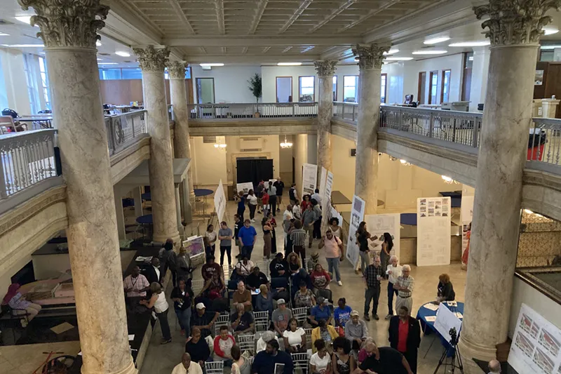 A high-angle view of a large, multi-story open room with marble columns, filled with people attending a community meeting.