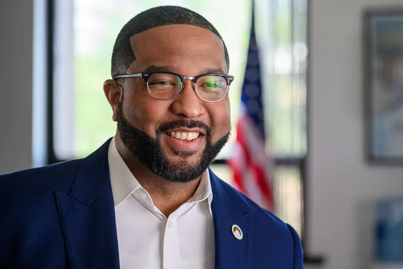 Professional headshot of Gary Mayor Eddie Melton, a smiling man in a blue suit and glasses, with an American flag visible behind him.