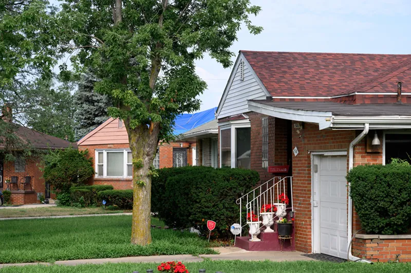 A residential neighborhood street view showing brick houses with hedges and red-shingled roofs on a bright green lawn.