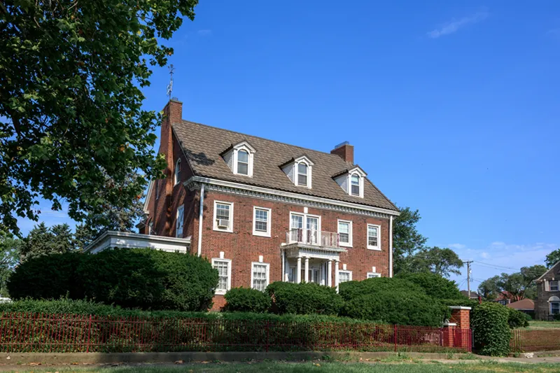 A two-story red brick house with a brown shingled roof and three dormers is surrounded by tall green hedges and a low red fence.